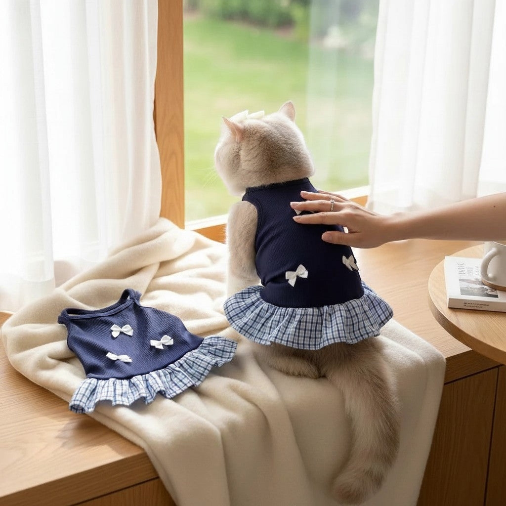 The Spoiled Pet Club blue polyester pet dress with white bows and a plaid ruffle skirt worn by a cat near a window.