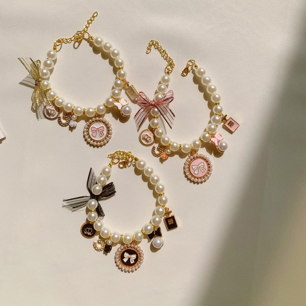 The Spoiled Pet Club pearl pet collars with bow and charm accents in pink, black, and white on a neutral background.
