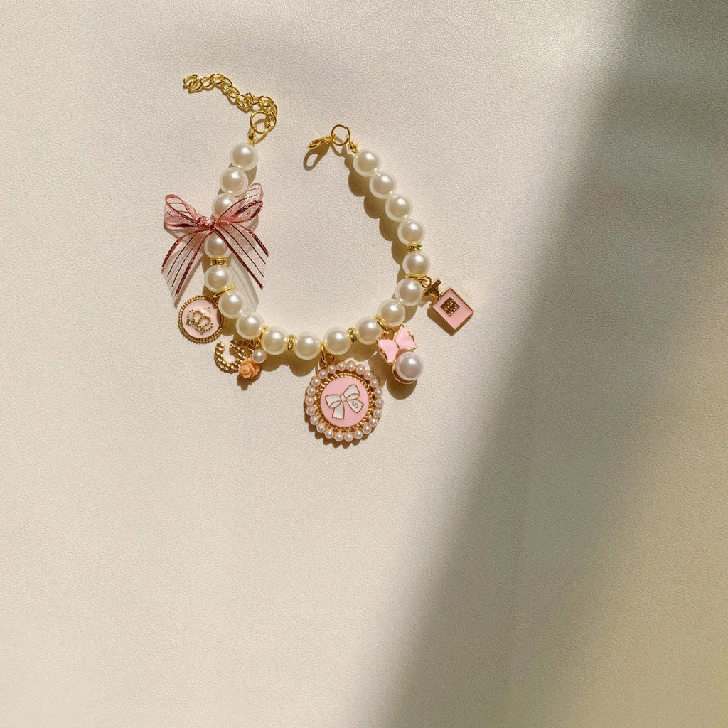 The Spoiled Pet Club pearl pet necklace with pink bow, charms, and bell accent on a cream background