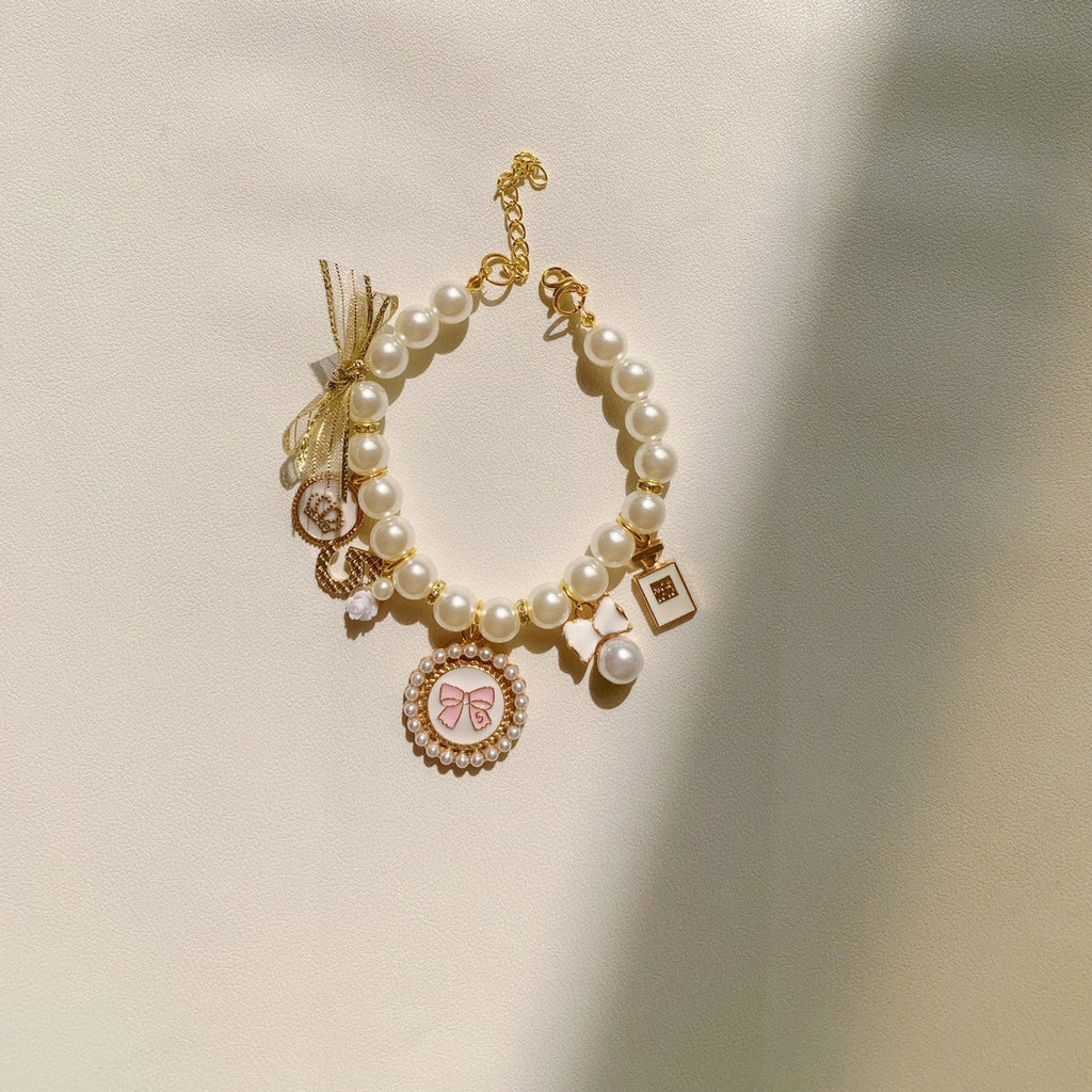 The Spoiled Pet Club pearl pet necklace with pink bow charm and gold accents on a cream background